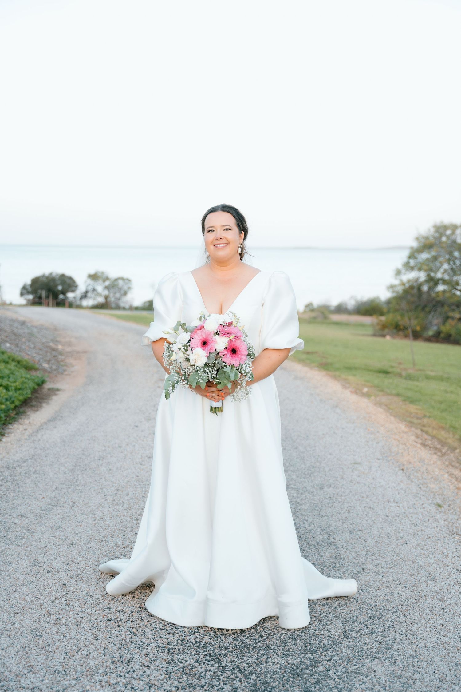 Bride in white A-line gown holding pink bouquet stands on gravel path with waterfront view behind her.