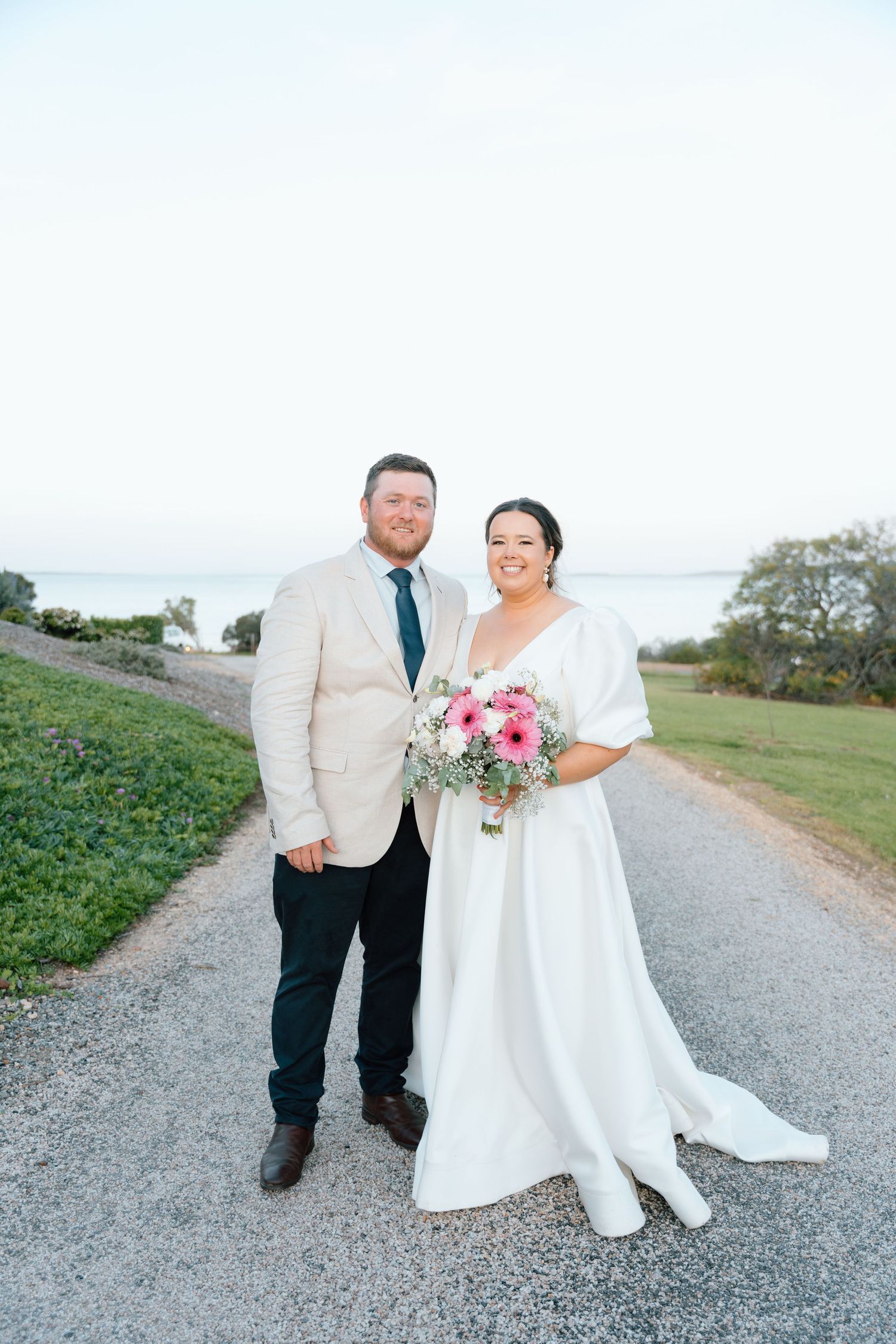 Bride and groom smiling on a gravel path by the water, she holds pink flowers in a white gown, he wears a tan suit.