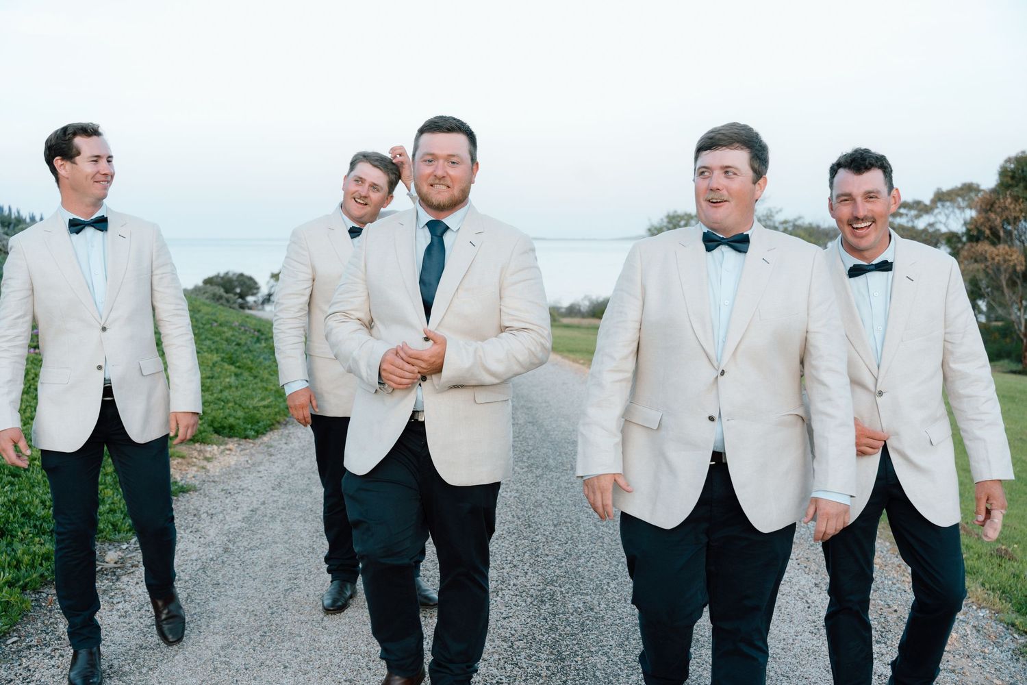 Five groomsmen in white tuxedo jackets and black bow ties walk together on a gravel path by the ocean.
