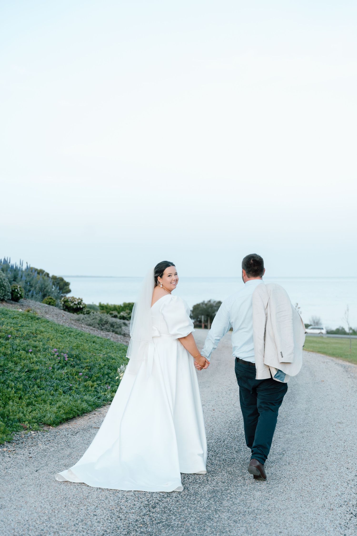 Bride in white gown and groom hold hands walking along a scenic coastal path at dusk after their wedding.