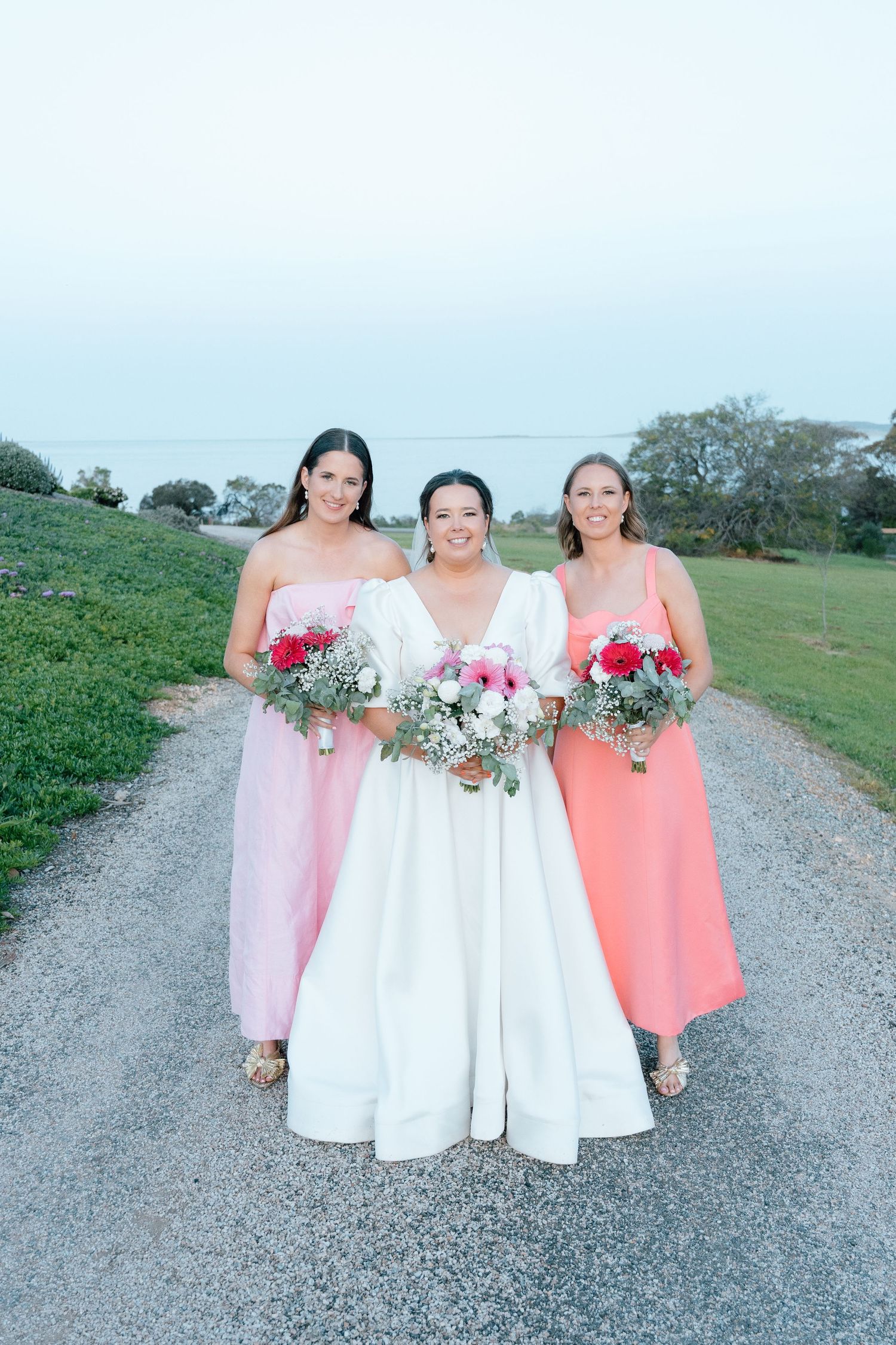 Bride in white gown walks with two bridesmaids in pink and coral dresses, holding bouquets on a gravel path.
