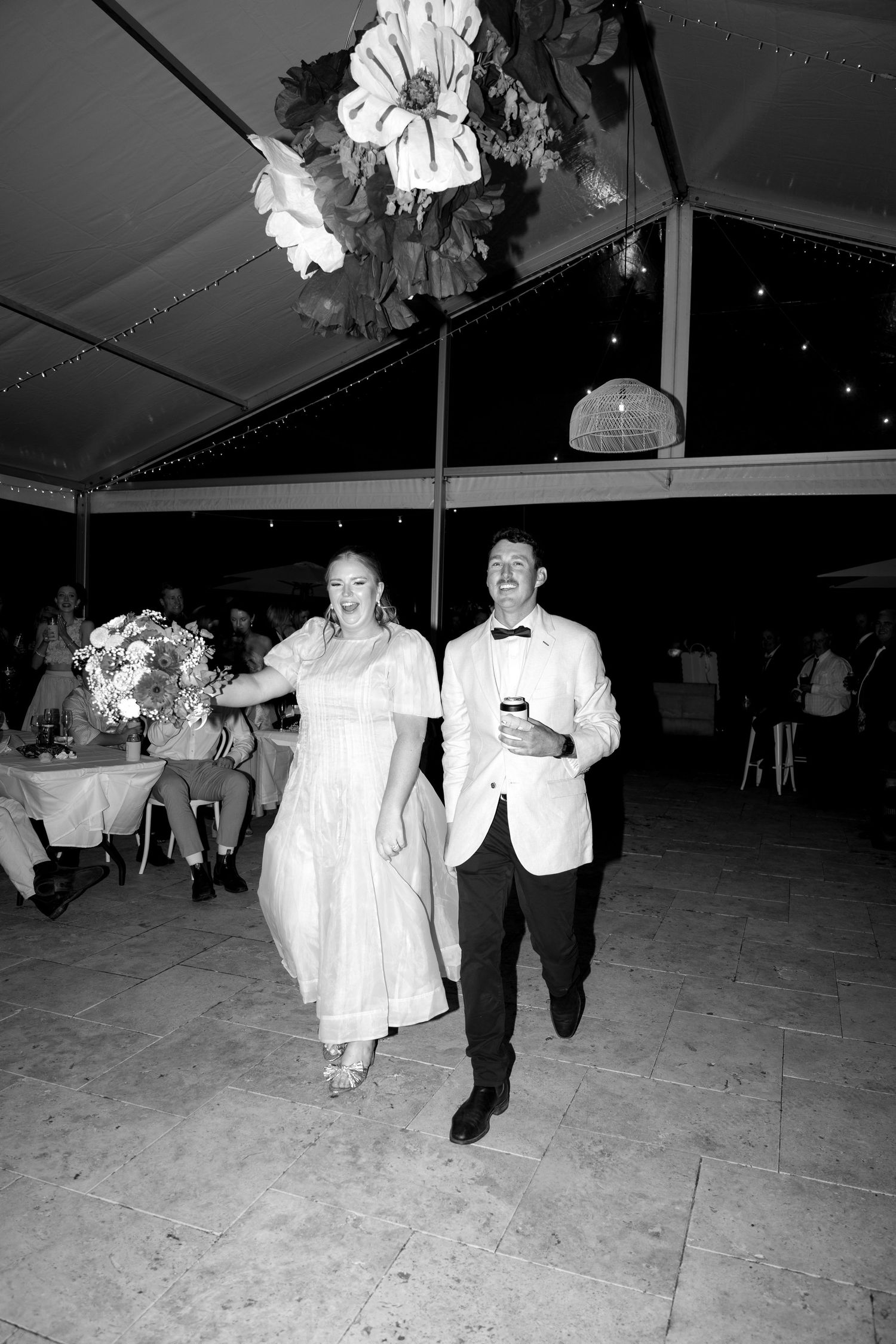 Black and white photo of a bride and groom walking hand in hand at their outdoor wedding reception under a tent.