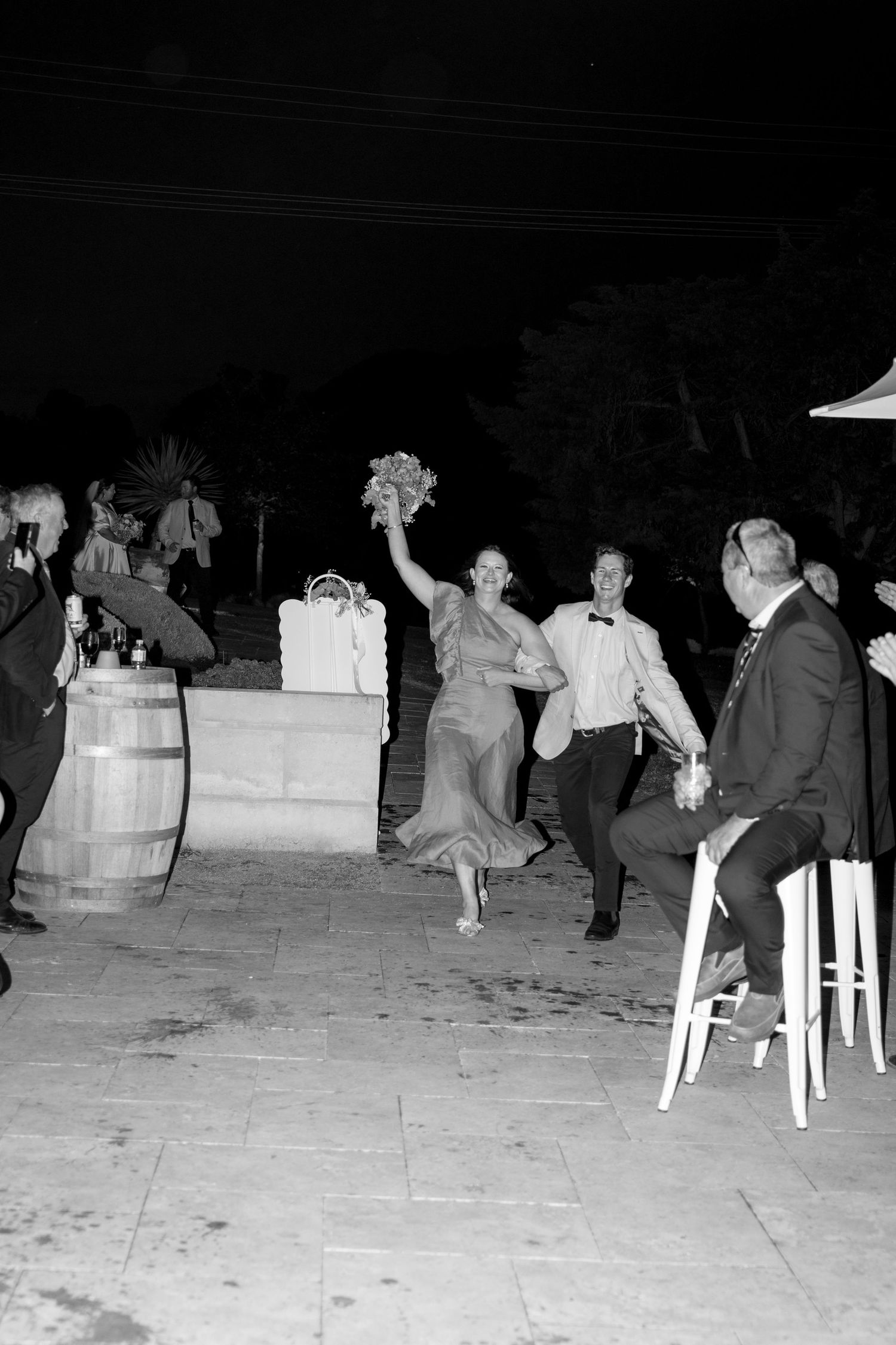 Black and white photo of a woman raising a bouquet while dancing with a man at an outdoor wedding reception at night.