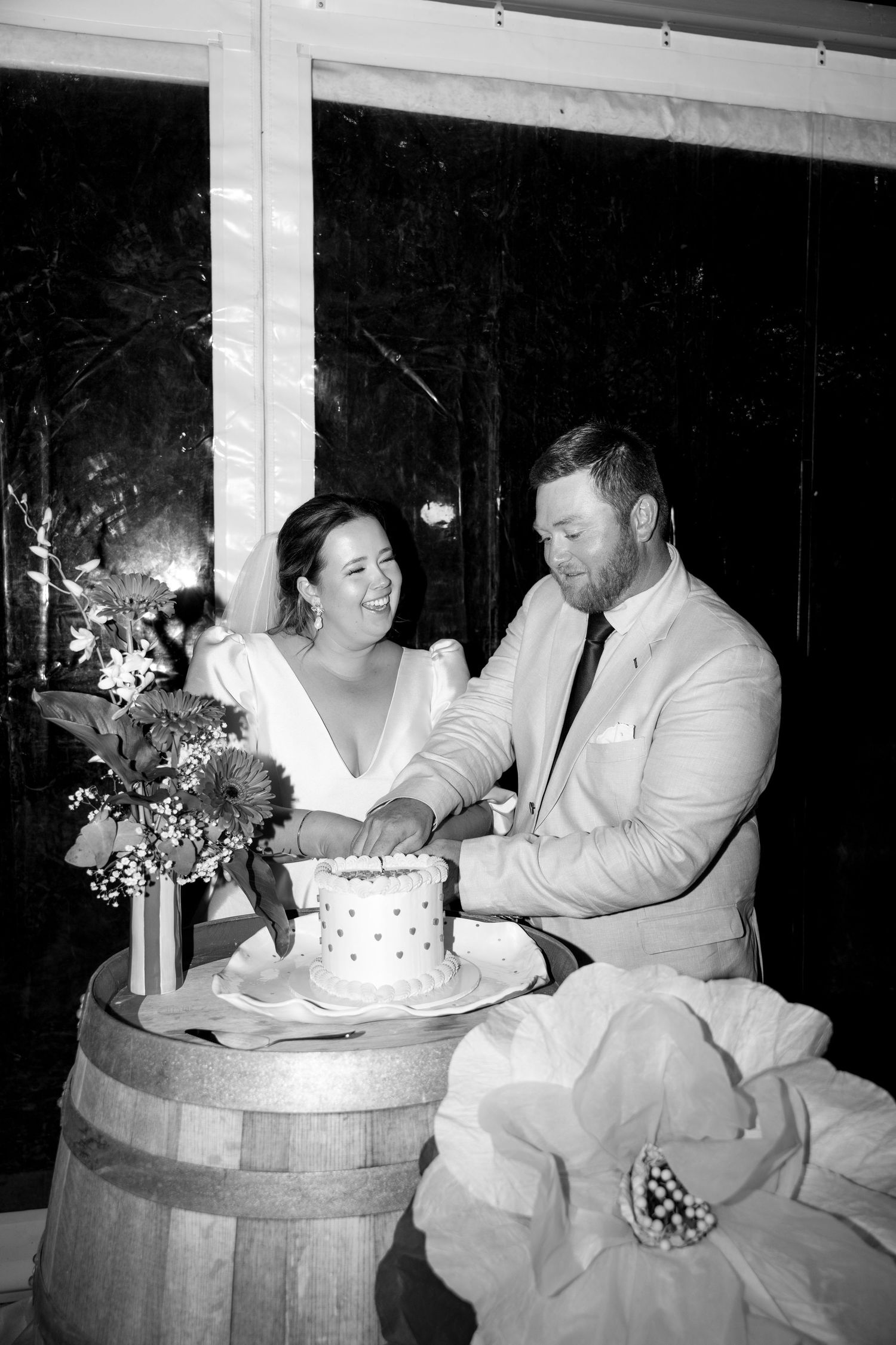 Bride and groom cut their wedding cake together on a wooden barrel at their reception.