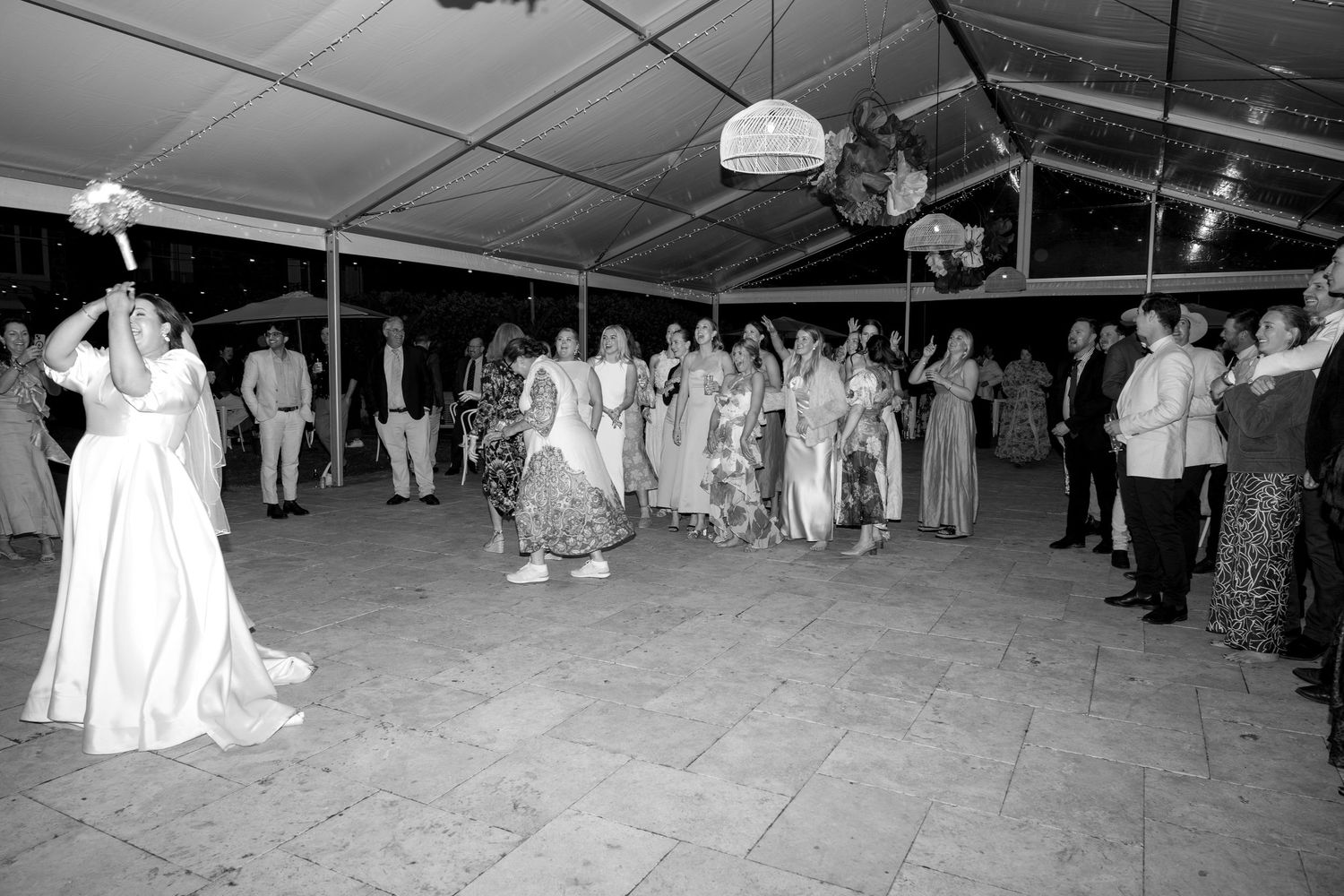 Bride tosses bouquet to guests at outdoor wedding reception under tent in black and white photo.