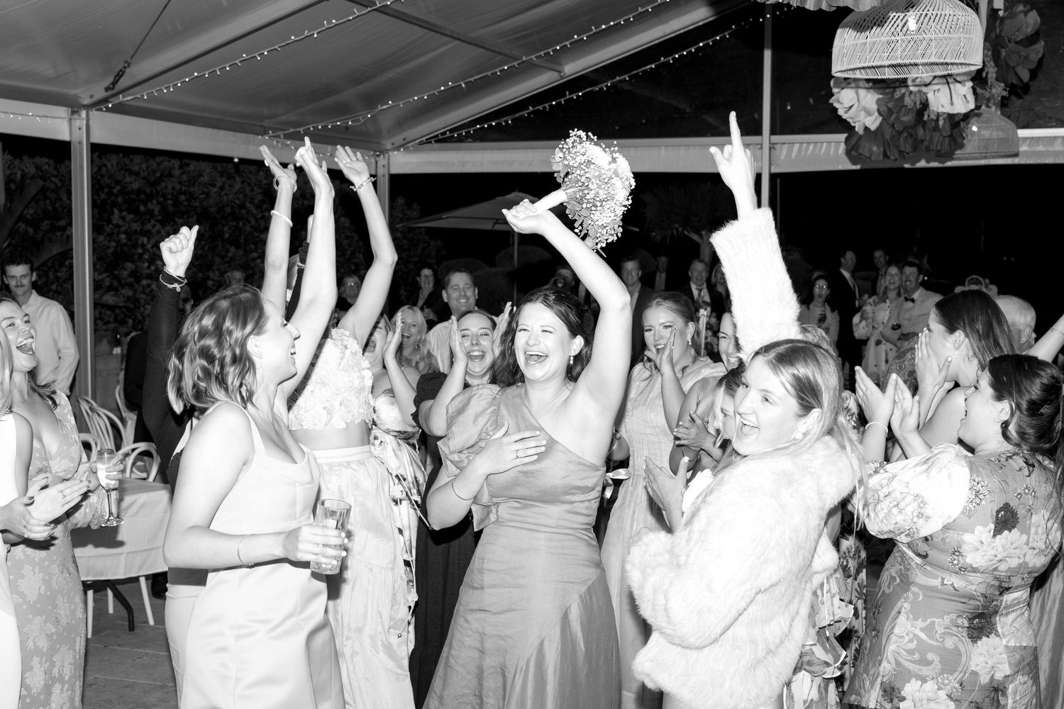 Excited wedding guests celebrate as the bride tosses her bouquet at an outdoor reception under a tent.