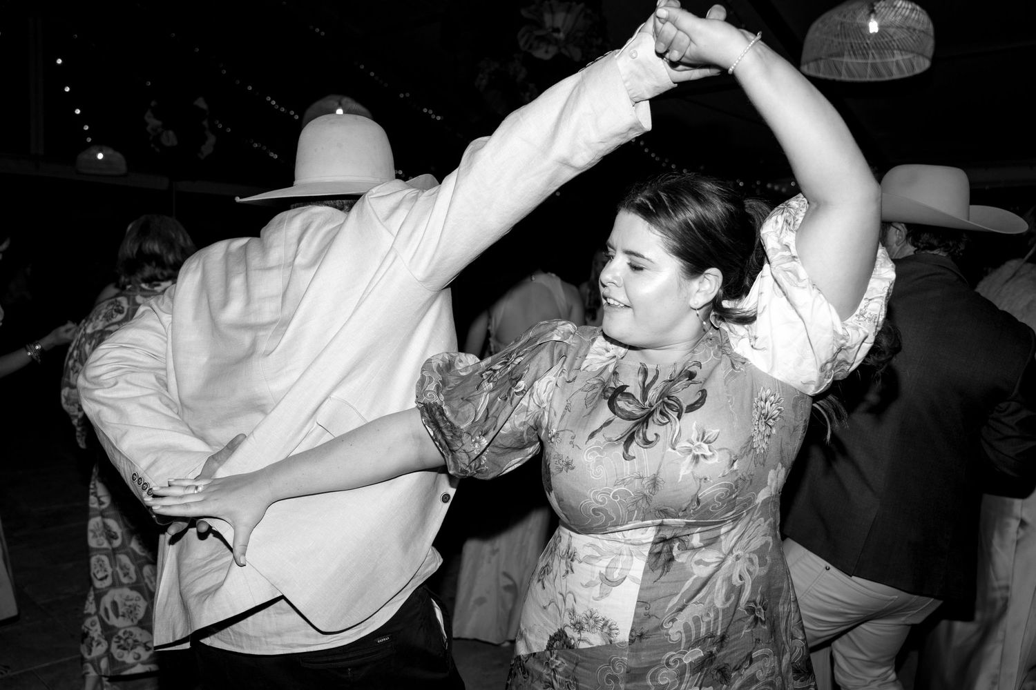 Couple dancing energetically at a party in black and white photo, woman in floral dress, man in cowboy hat.
