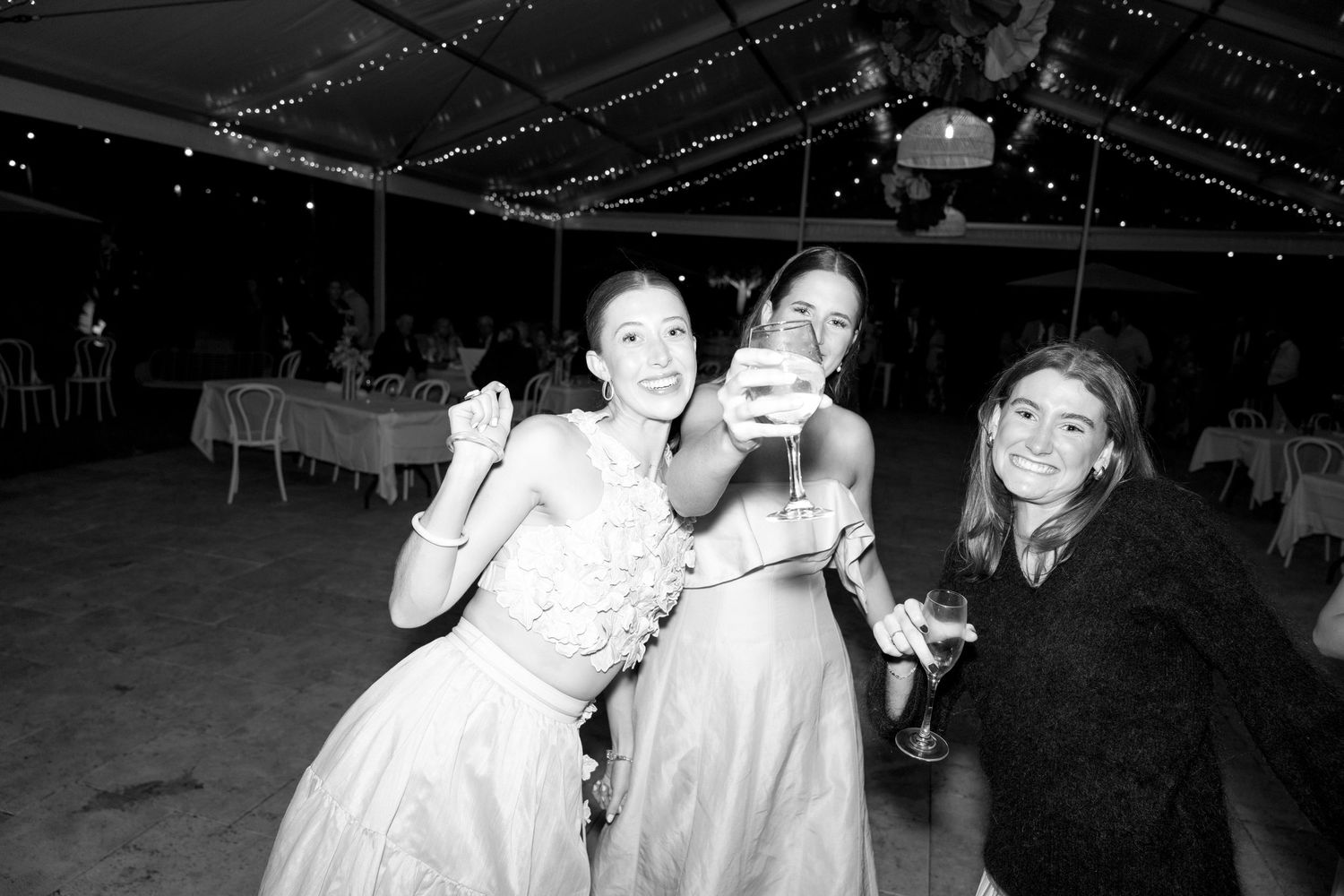 Three women in formal dresses celebrating at a wedding reception under a tent with string lights.