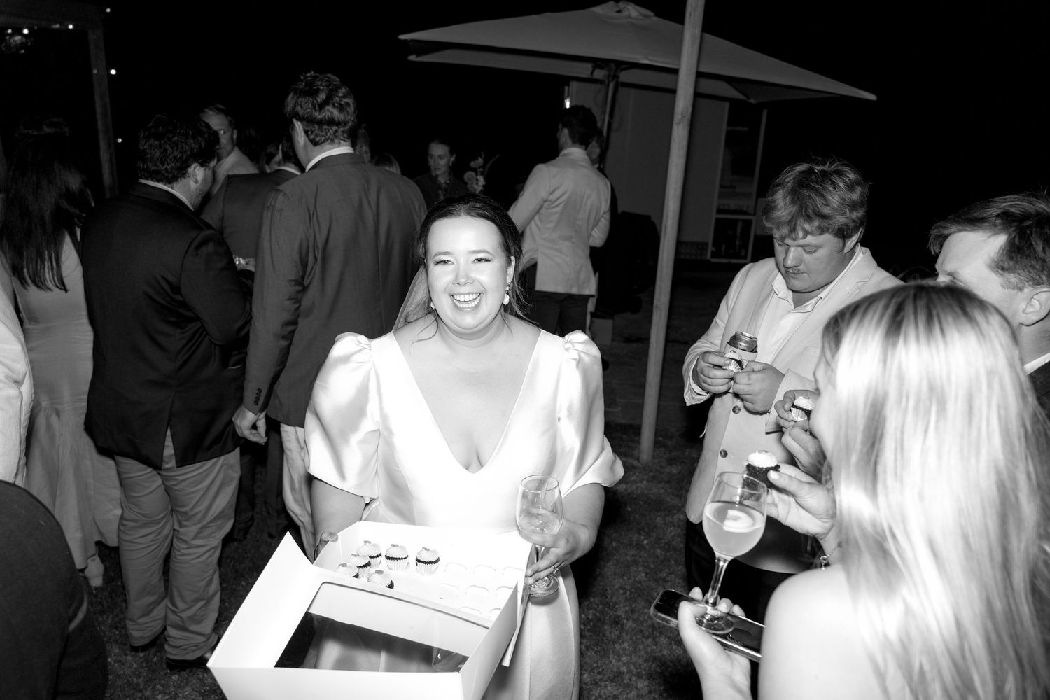 Smiling bride in white gown holding a box at an outdoor wedding reception with guests in the background.