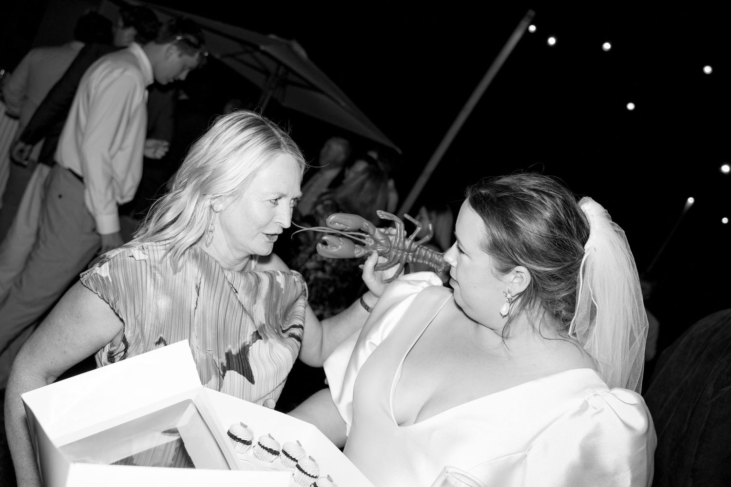 Bride in white gown laughs with a blonde guest while a violinist performs at the wedding reception.