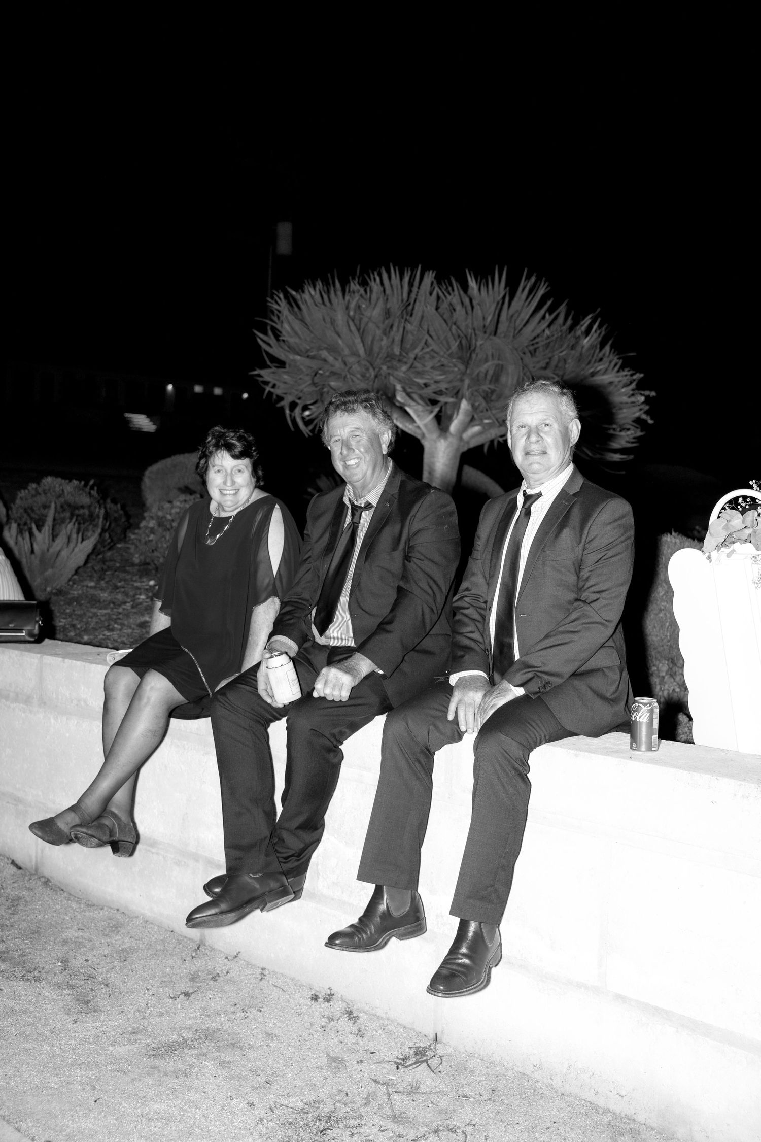 Three formally dressed wedding guests sit together on a white wall outdoors at night.
