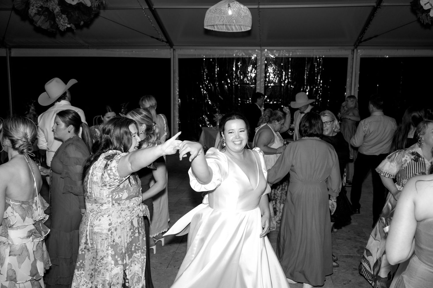 Bride in white wedding gown dances joyfully with guests on a crowded wedding reception dance floor.