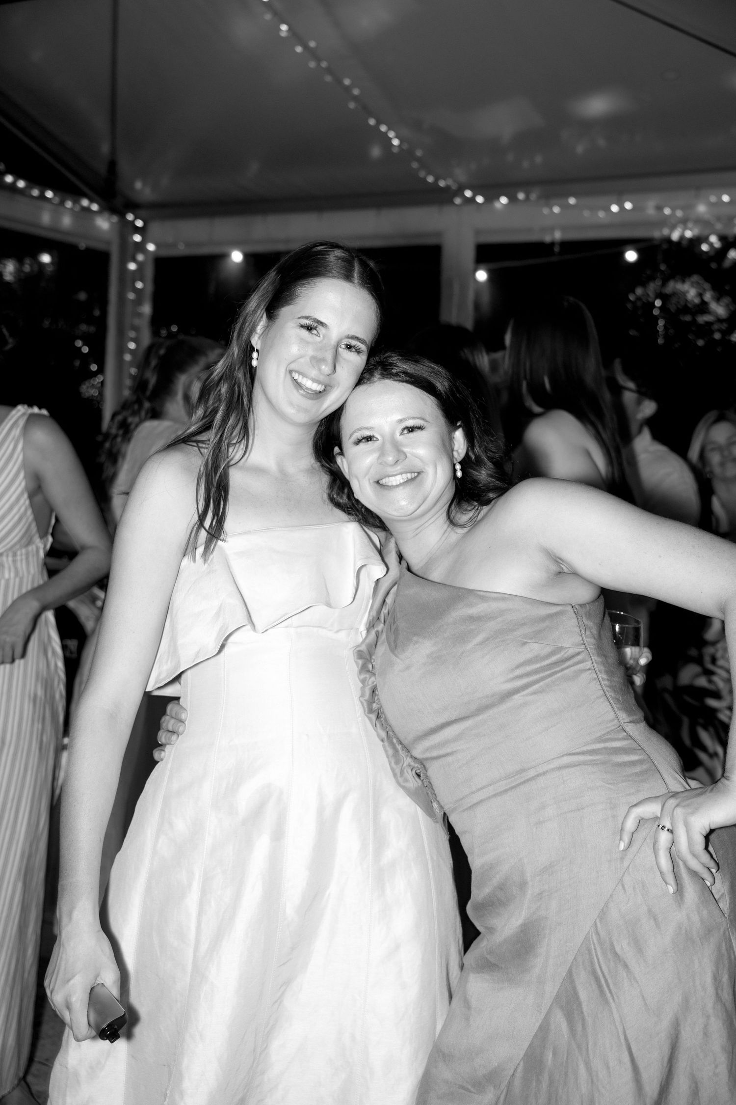 Two women in formal gowns smiling together at a wedding reception dance floor in black and white photo.