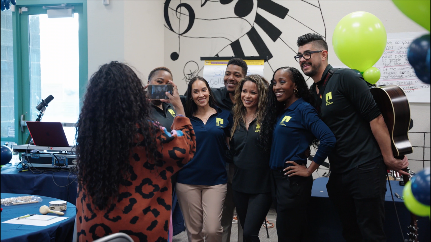 Group of young professionals in matching navy shirts posing for a photo at a music-themed event with balloons.