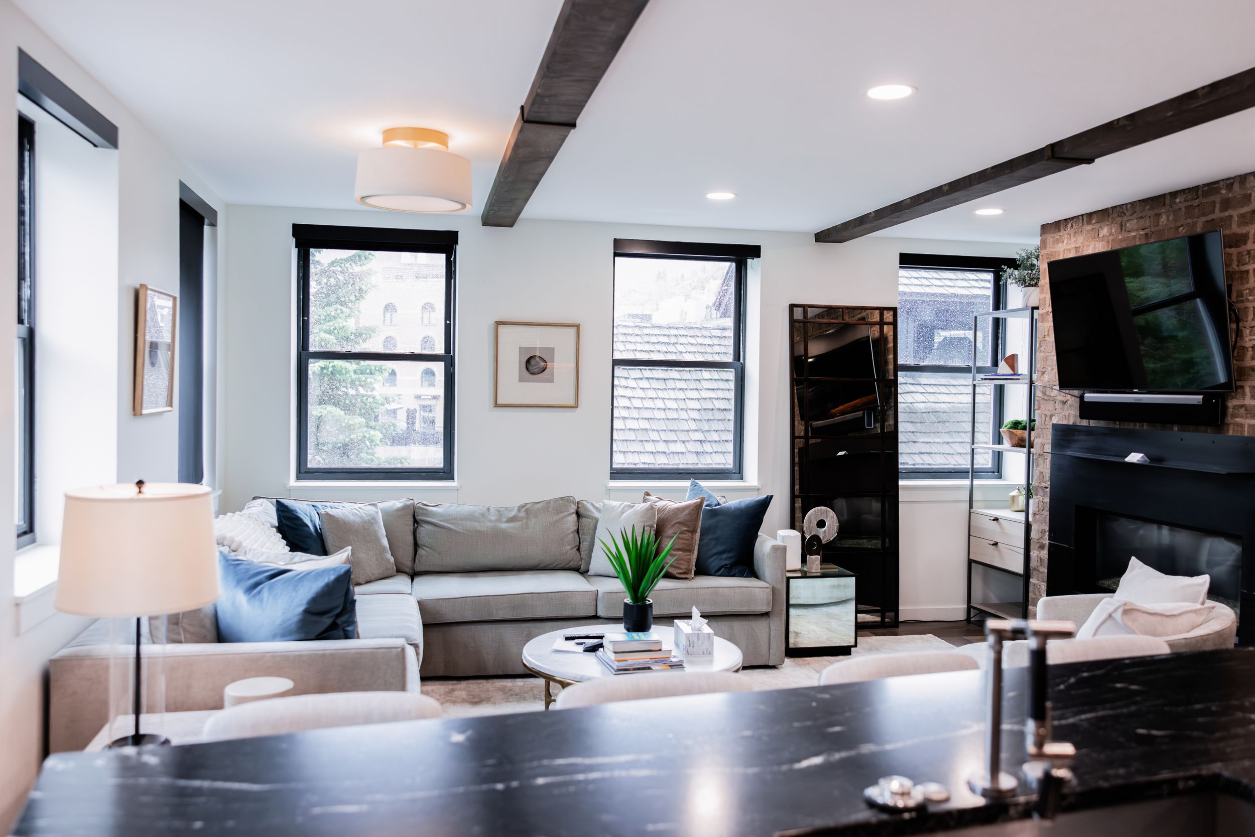 Bright modern living room with gray sectional sofa, exposed dark wood beams, mounted TV, black marble countertop and large windows.
