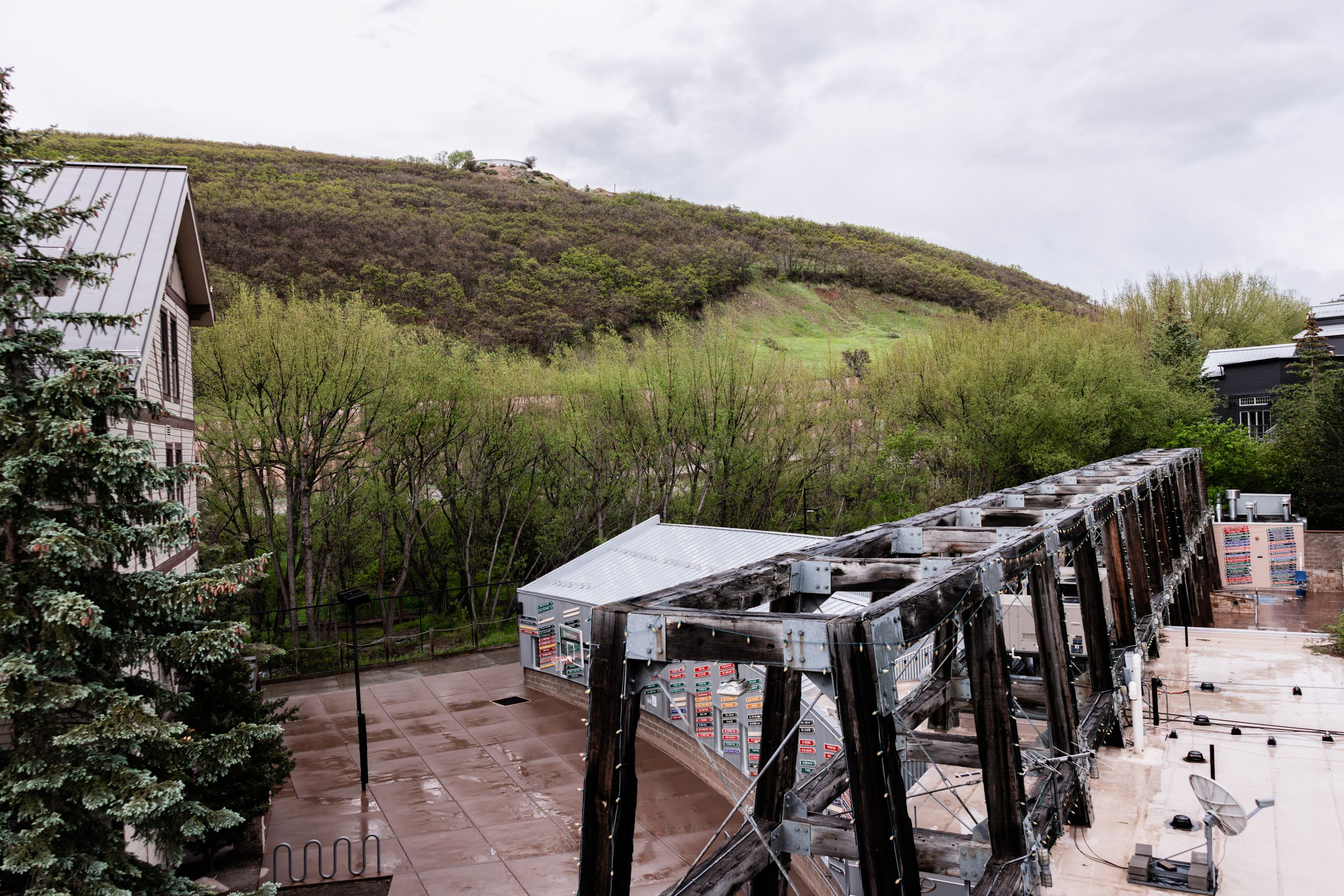 Black steel truss bridge spanning a river with green trees, a grassy hillside, and mountain-town buildings under a cloudy sky.