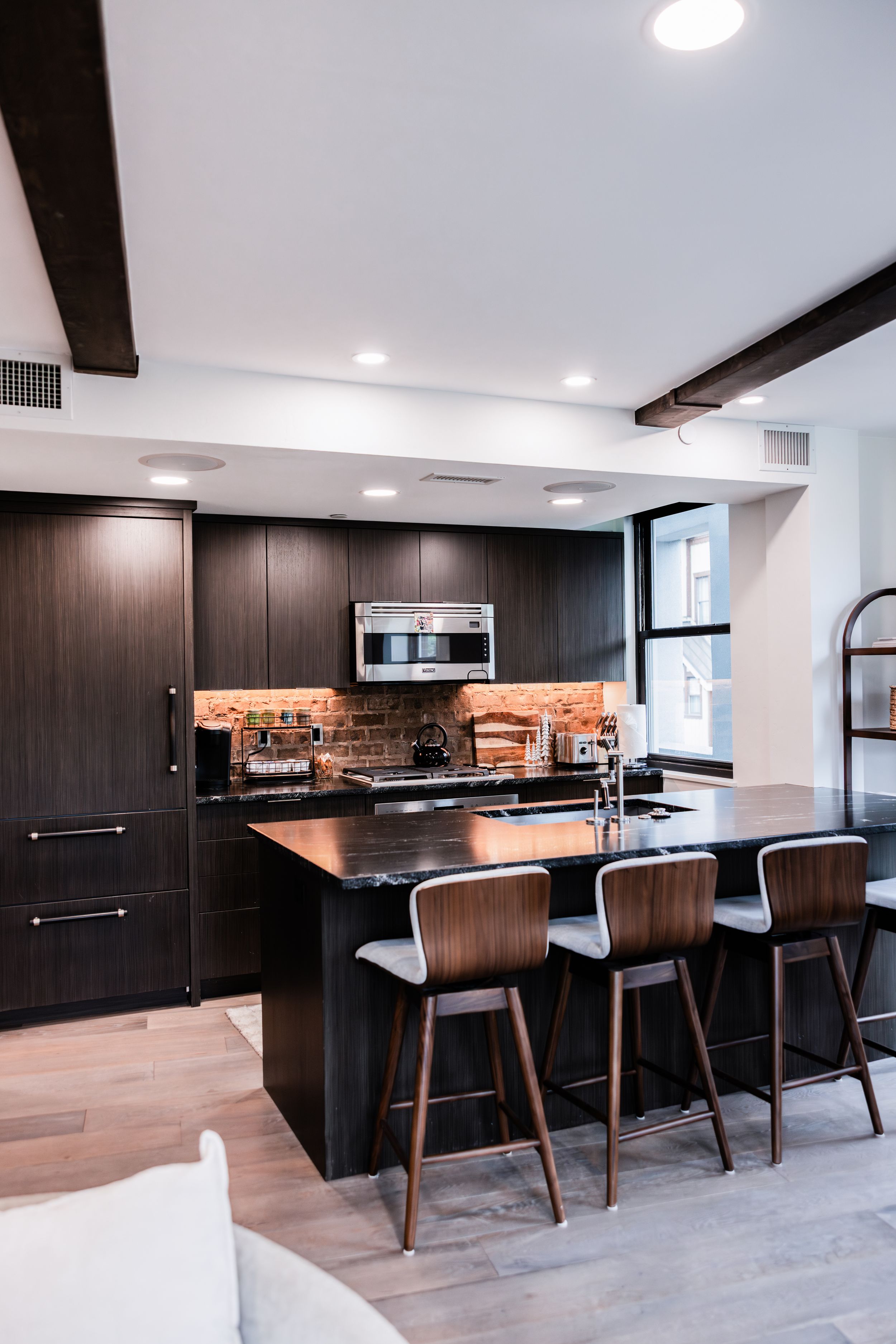 Modern kitchen with dark wood cabinets, brick backsplash, black granite island, and three leather bar stools beneath exposed ceiling beams.