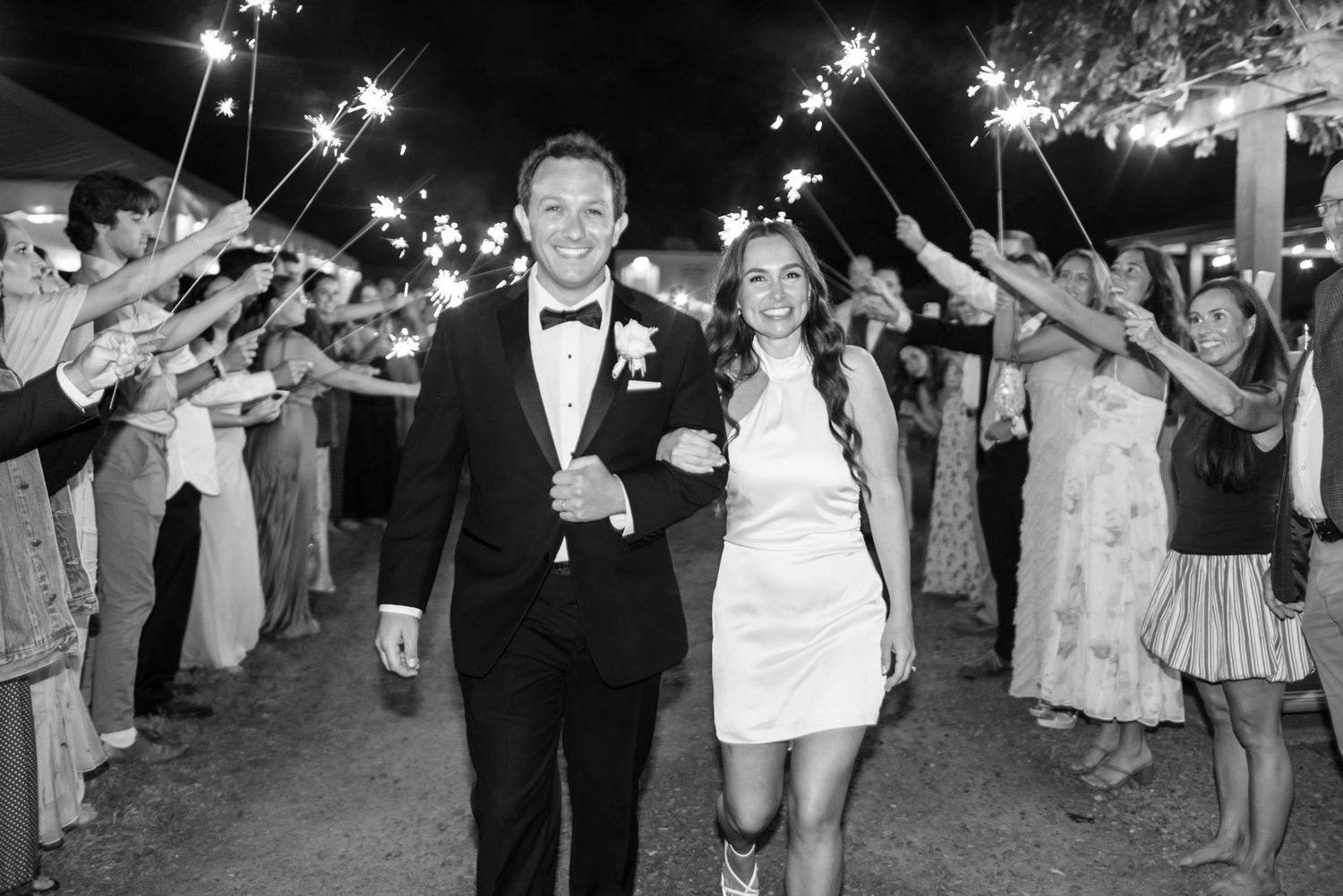 Bride in short white dress and groom in black tuxedo walk arm in arm as wedding guests hold sparklers during a festive evening send-off.
