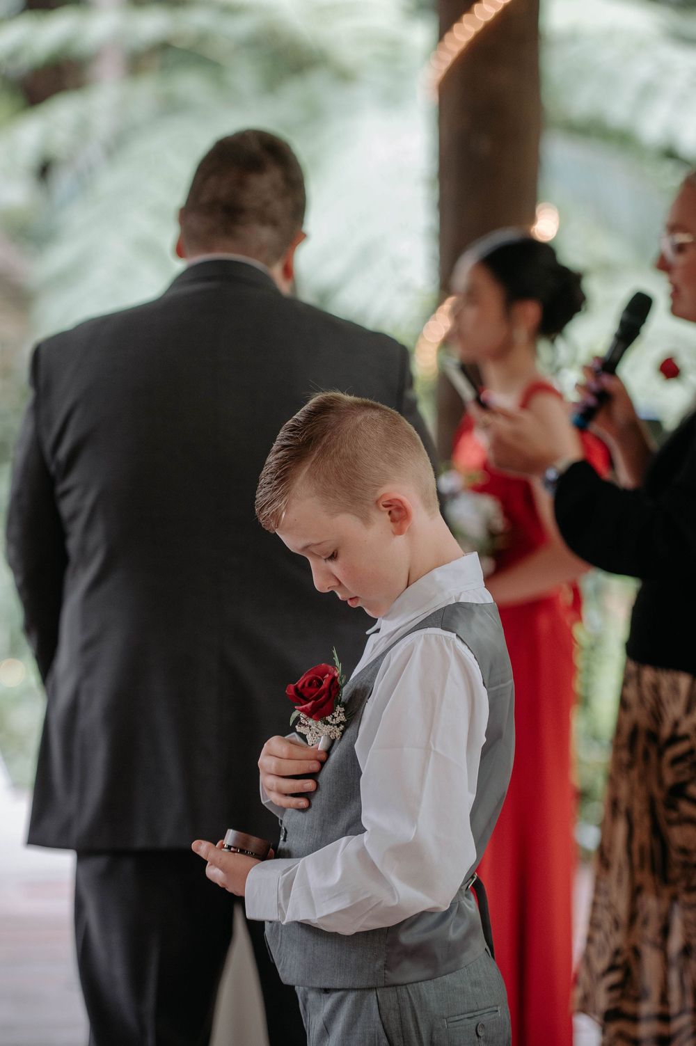 Young boy in a gray vest with a red rose boutonniere looks down holding a ring box during an outdoor forest wedding ceremony.