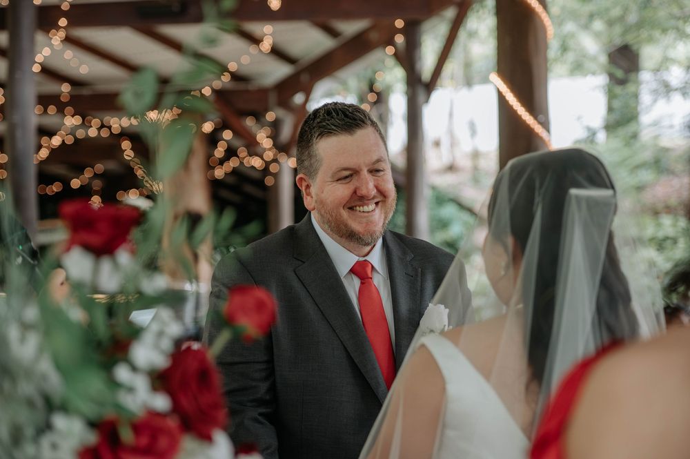 Smiling groom in a dark suit and red tie looks at his veiled bride during an outdoor wedding ceremony with red floral arrangements.