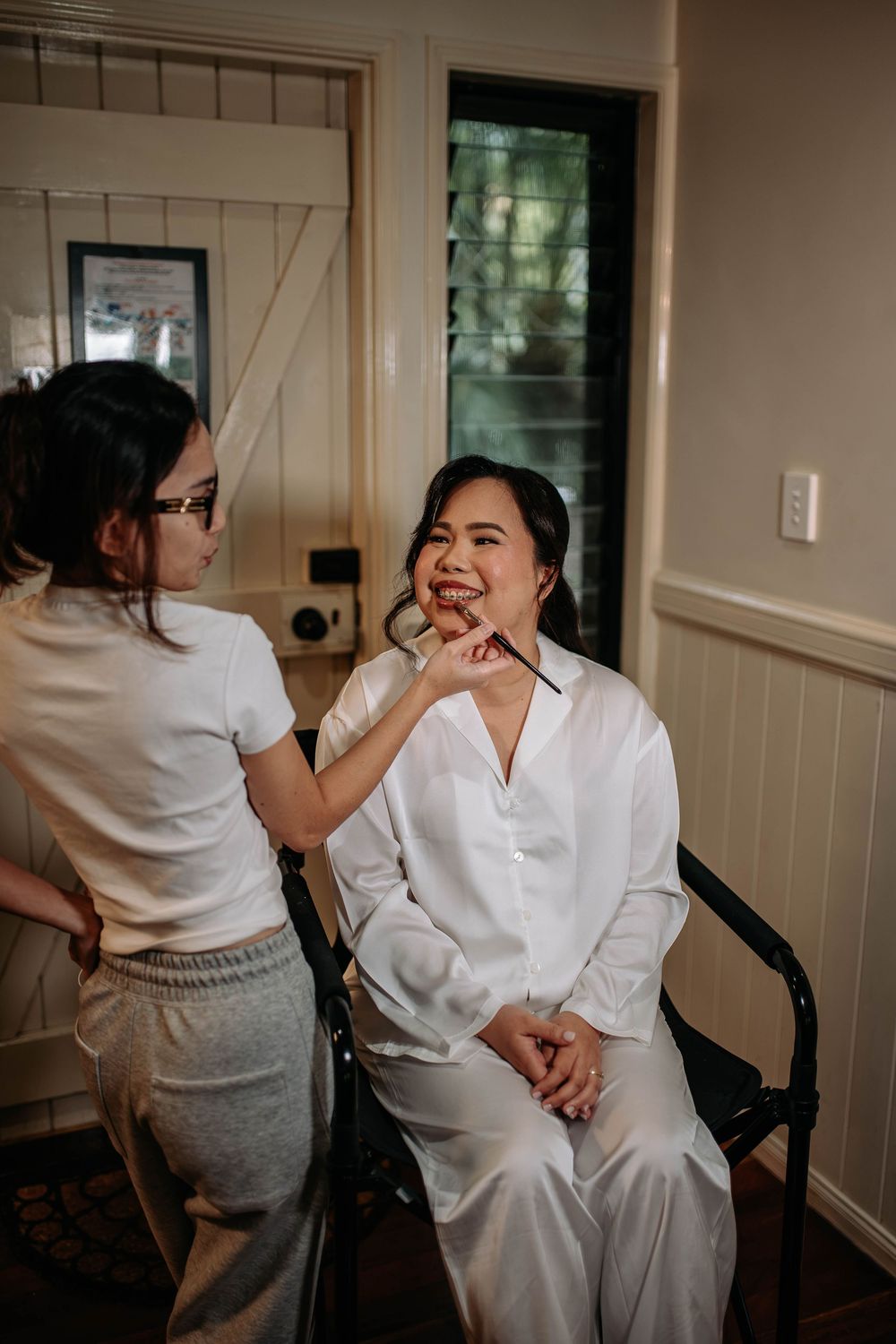 A makeup artist applies lipstick to a smiling woman in a white satin robe seated in a chair, getting ready for a wedding in a bright room.
