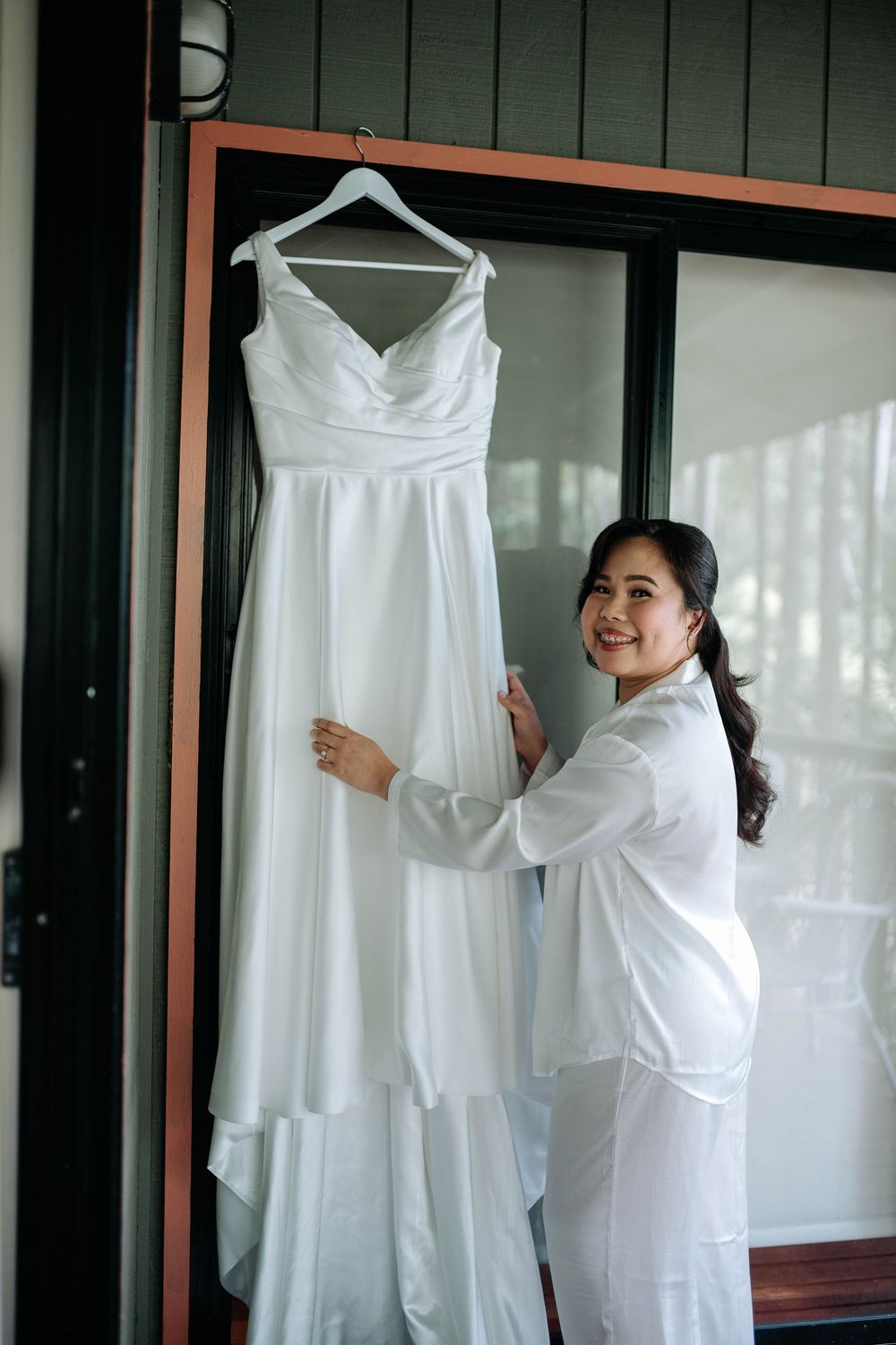 Smiling bride in a white satin robe gently touches her elegant white wedding gown hanging on a window frame before the ceremony.
