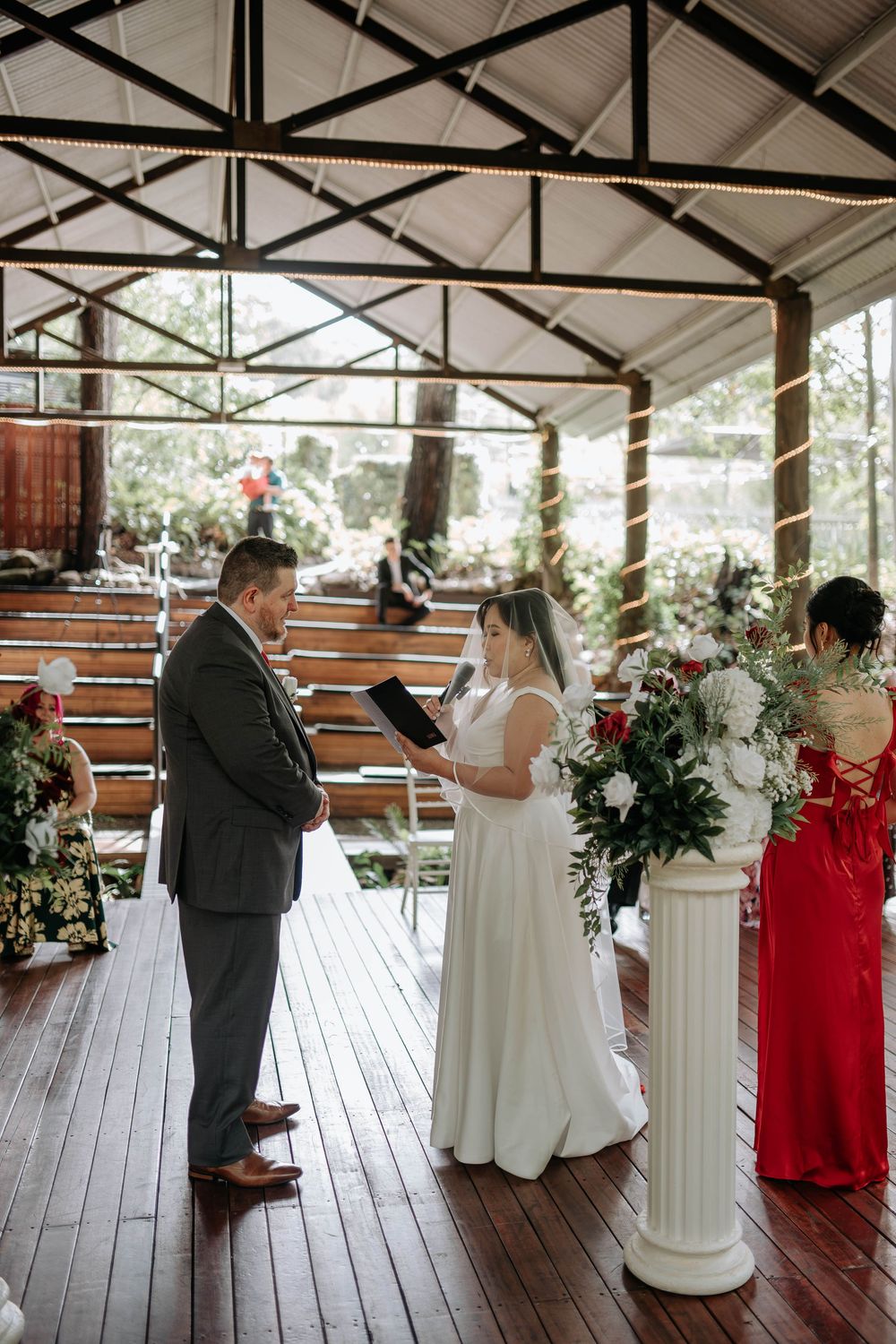 Bride in a white gown reads vows to a groom in a gray suit during an outdoor wedding ceremony on a wooden deck with floral arrangements.