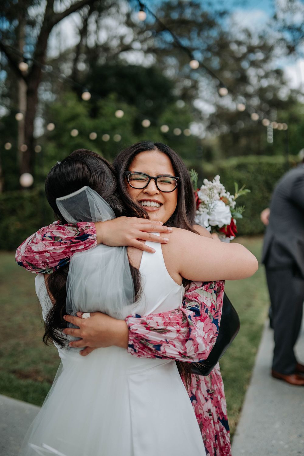 Smiling guest in a floral dress and glasses hugs a bride in a white gown and veil at an outdoor wedding with string lights.
