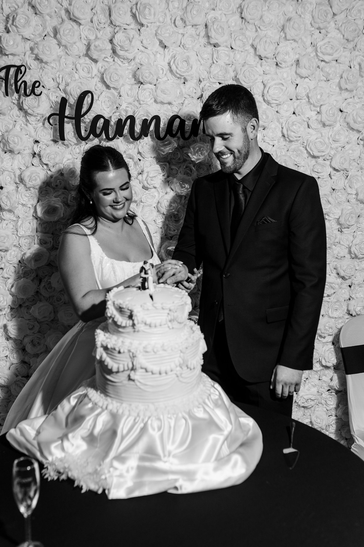 Smiling bride and groom cutting their tiered wedding cake in front of a white flower wall in a black and white photo.