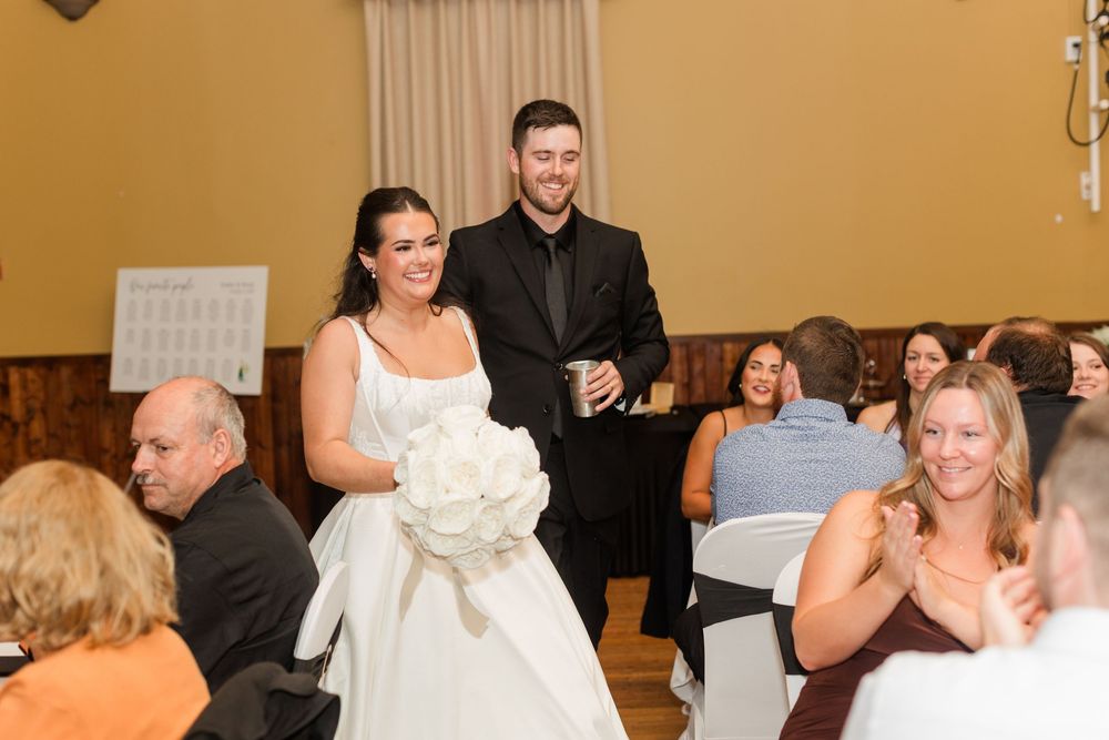 Bride in a white gown holding a white bouquet walks beside a groom in a black suit as smiling wedding guests applaud at the reception.