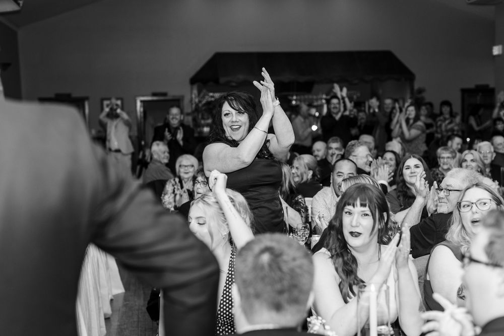 Cheering wedding guests clap and celebrate in a crowded reception hall, with a smiling woman standing and applauding in the center.