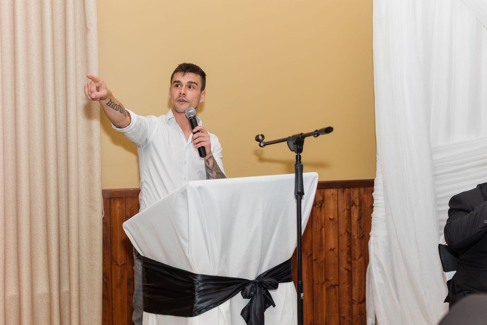 Man in a white shirt speaks into a microphone while pointing outward beside a podium draped in white cloth with a black bow.