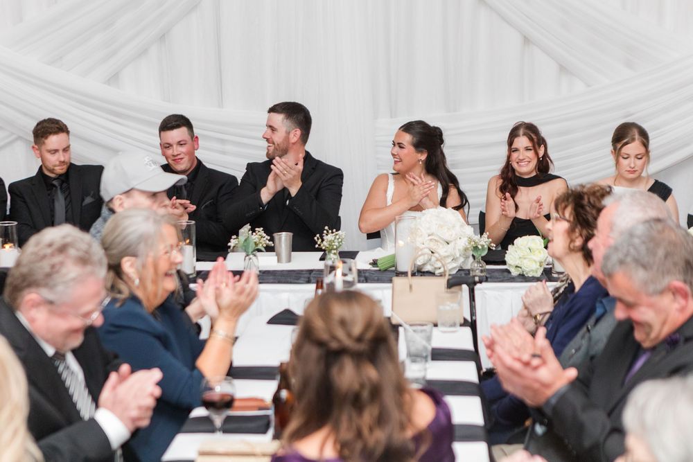 Wedding guests in formal attire smile and applaud at a long head table decorated with white floral centerpieces and draped white fabric.