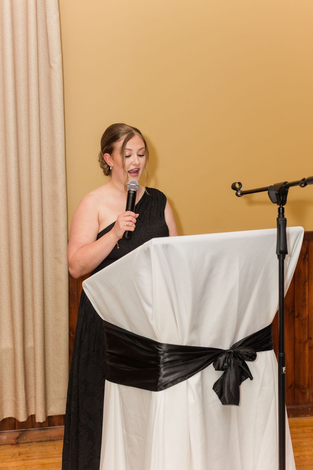 Woman in a black one-shoulder dress speaks into a microphone beside a podium draped in white linen with a black satin bow.
