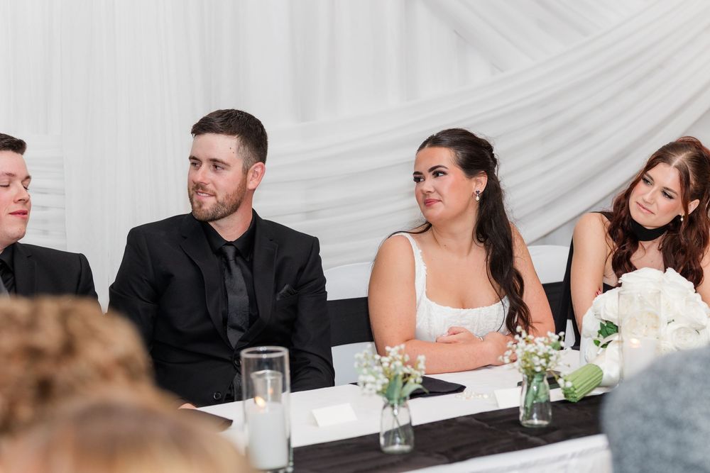 Wedding guests in formal attire sit at a decorated head table with white floral centerpieces, smiling and looking toward the room.