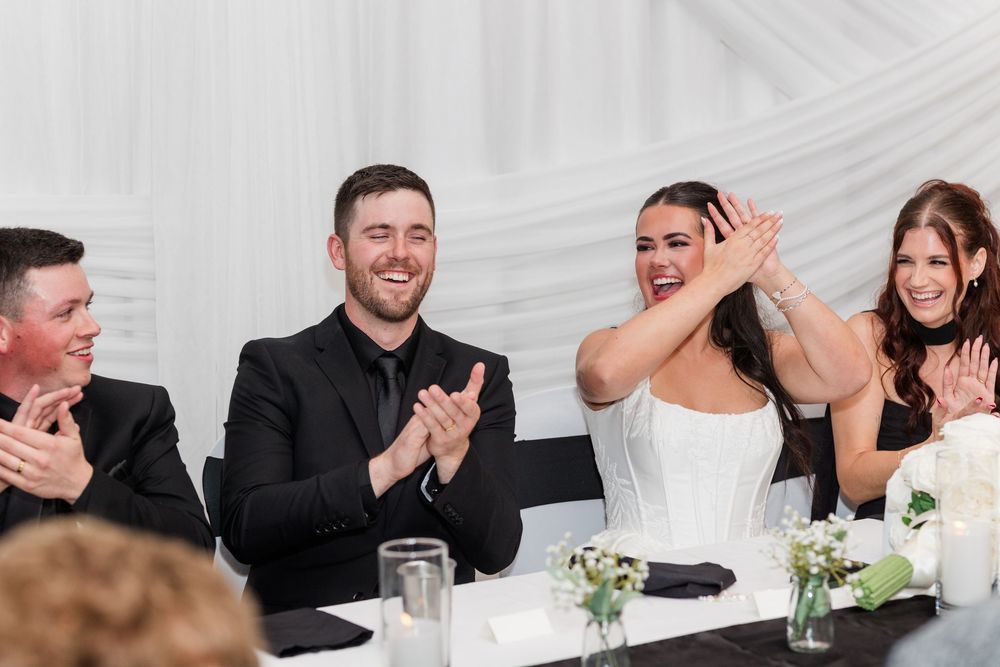 Bride in white dress and wedding guests in formal attire smile and applaud at a reception table decorated with flowers and candles.