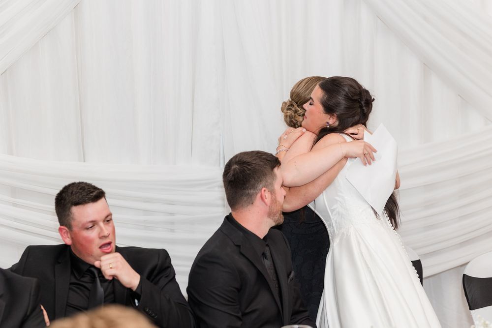 A bride in a white gown hugs a guest warmly while two men in dark suits sit nearby at a wedding reception with draped white fabric.