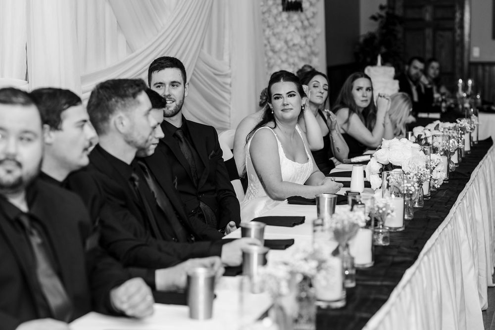 Black and white photo of wedding guests in formal attire seated along a long reception head table decorated with flowers and candles.