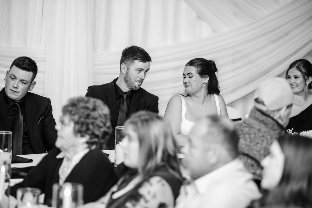 Black and white photo of wedding guests seated at a banquet table, with two men and two women smiling and looking off to the side.