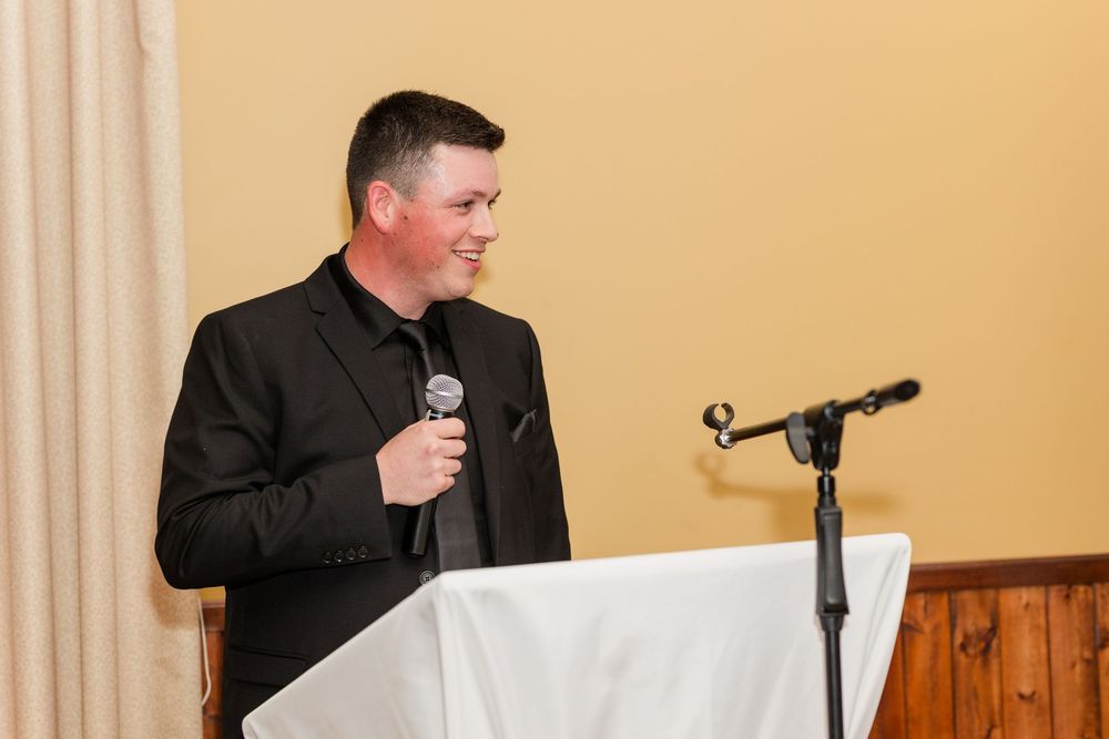 Man in a black suit smiles while holding a microphone and speaking at a white-draped podium with a second microphone stand nearby.