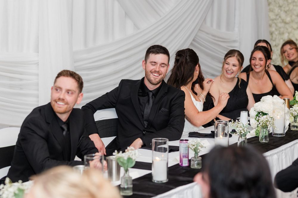 Smiling groomsmen in black suits and bridesmaids in black dresses sit at a wedding head table with white flowers and candles.
