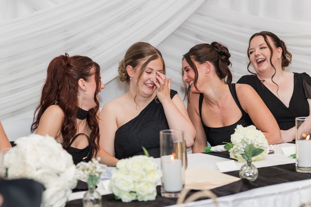 Five bridesmaids in black dresses laugh together at a wedding reception table decorated with white flowers and candles.