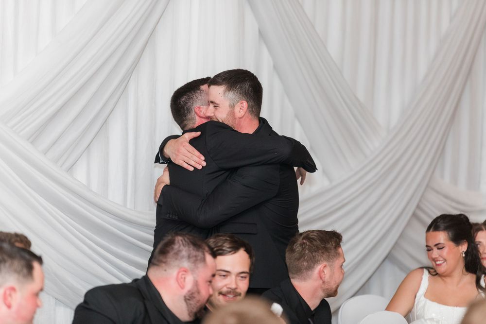 Two men in dark suits share a warm embrace at a wedding reception while seated guests smile and look on beside draped white fabric.