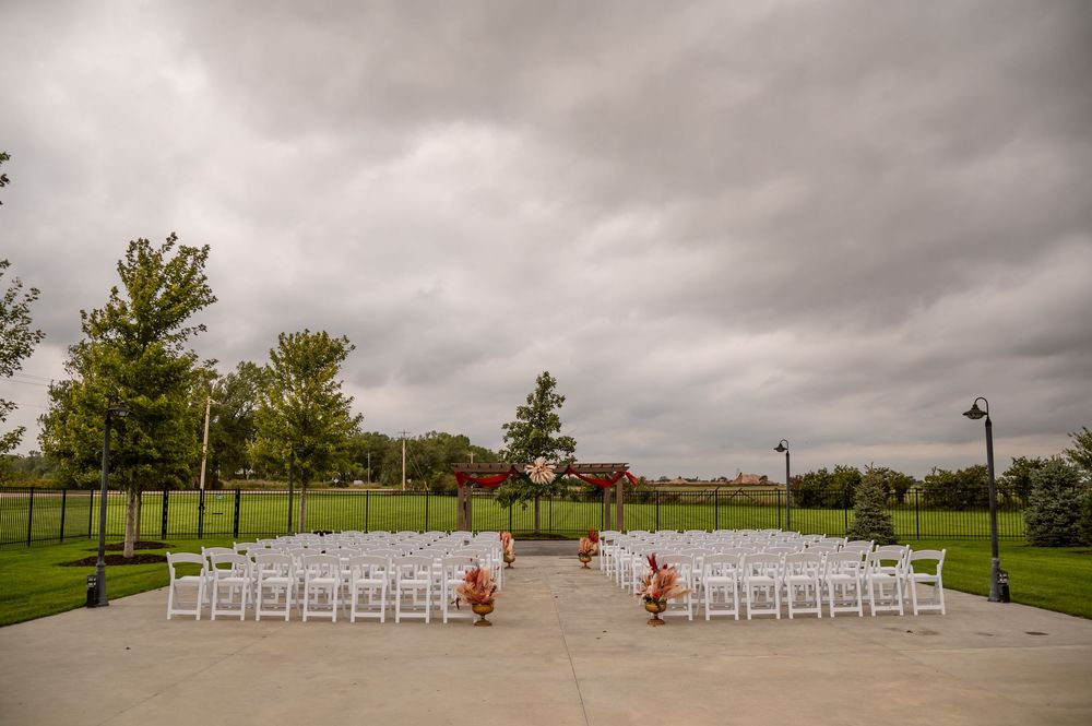Outdoor wedding ceremony setup with white chairs, a floral arch, and orange flower arrangements on a patio under a cloudy sky.