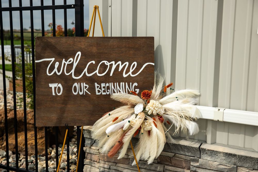 Rustic wooden welcome sign reading Welcome to Our Beginning beside a dried pampas grass and floral arrangement at a wedding entrance.