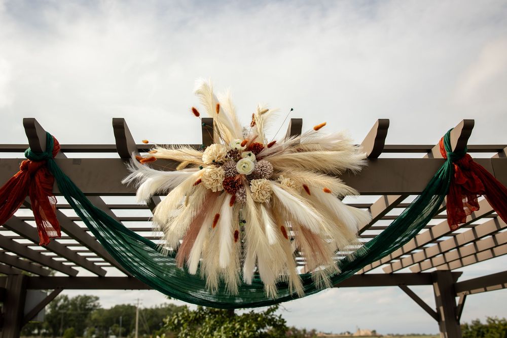 Boho wedding arch with pampas grass, dried flowers, and teal and rust draped fabric on a wooden pergola against a bright cloudy sky.
