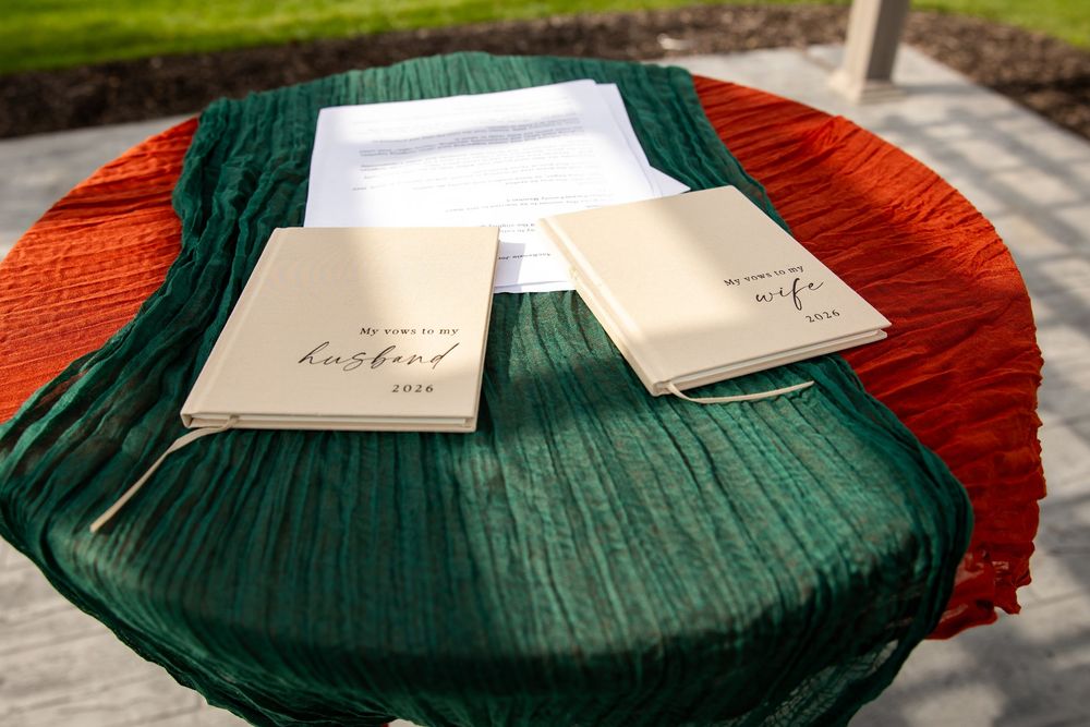Two cream wedding vow books labeled husband and wife rest on a green table runner over an orange cloth beside printed ceremony papers.