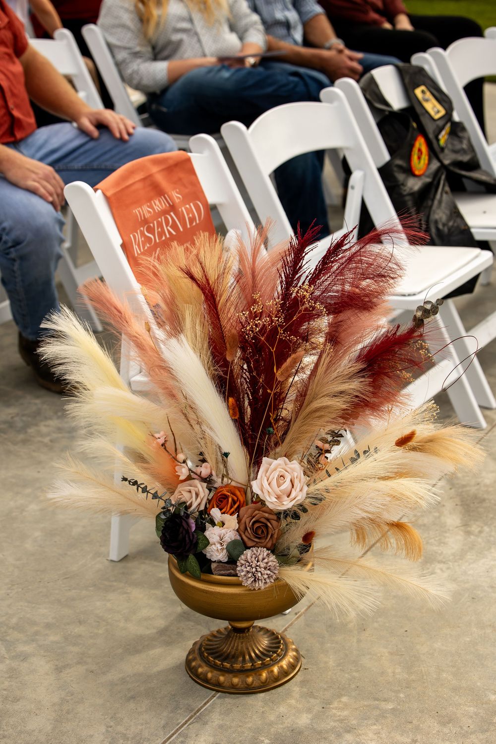 Boho pampas grass and dried floral arrangement in a gold pedestal vase beside white folding chairs with a reserved sign and seated guests.