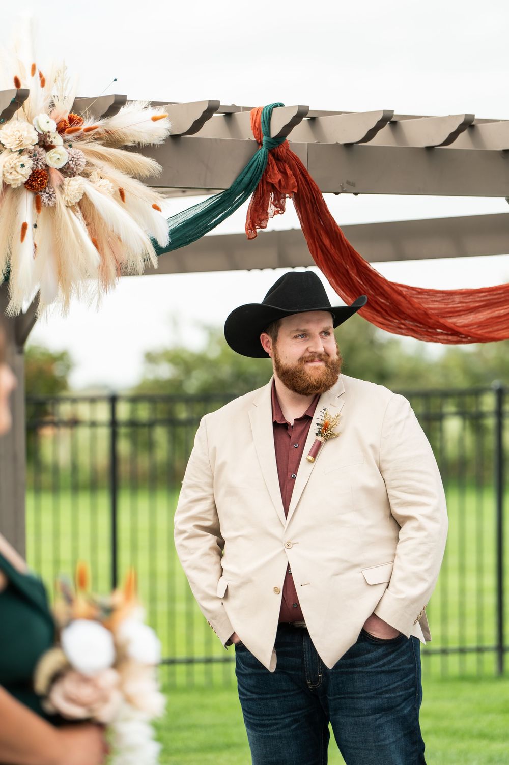 Bearded groom in a black cowboy hat and cream blazer stands under a wedding pergola decorated with pampas grass and rust and teal draping.
