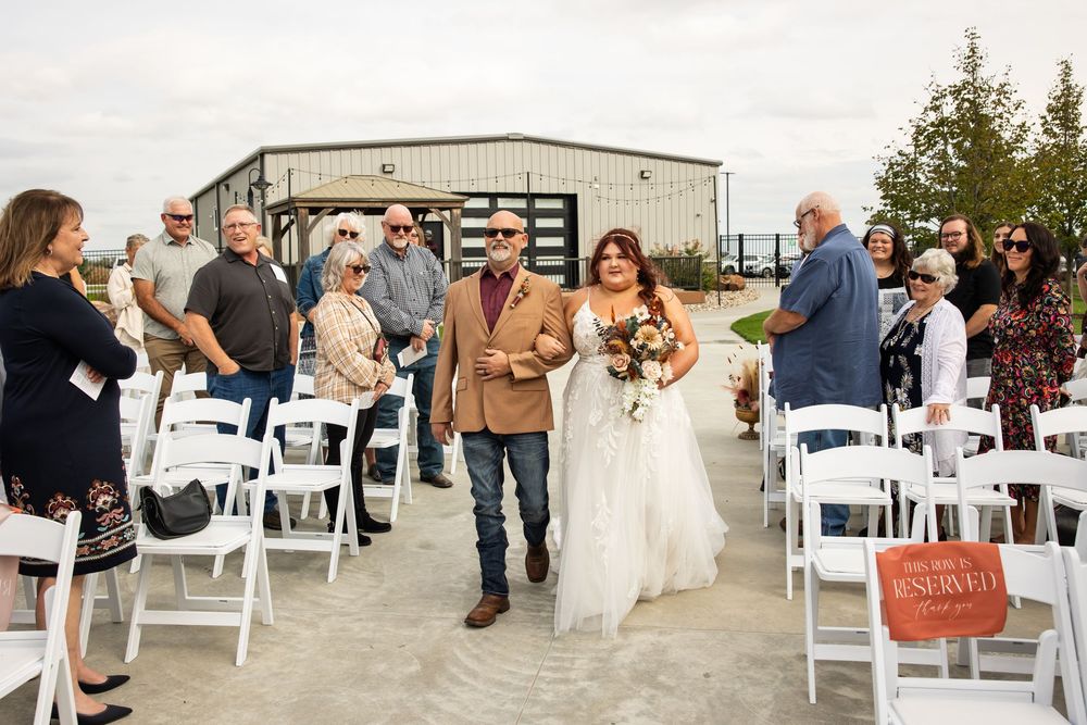 Bride in a white gown holding a bouquet walks down the aisle with a man in a tan blazer as guests watch at an outdoor wedding ceremony.